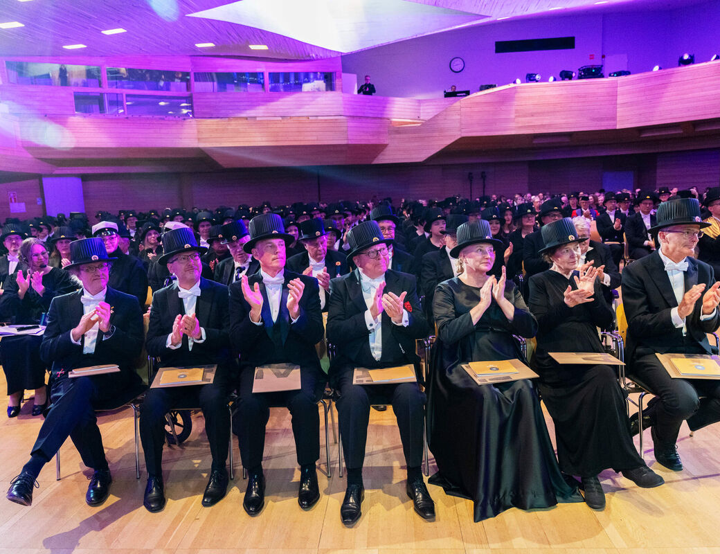 Audience in formal black attire and hats applaud in a large, purple-lit wooden hall