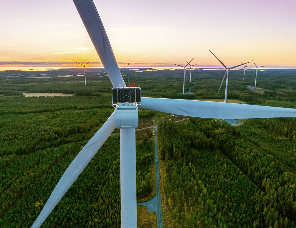 Close view of a wind turbine above a green forest, with more turbines and a sunset sky in the distance