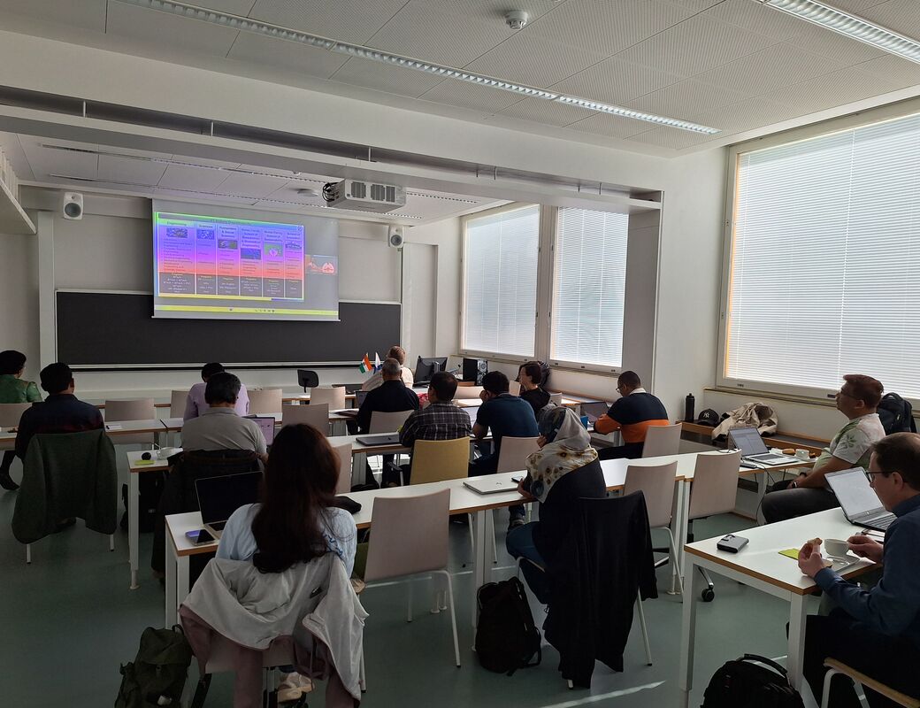 Students sit at desks with laptops in a bright classroom, watching a colourful slide on a projector screen.