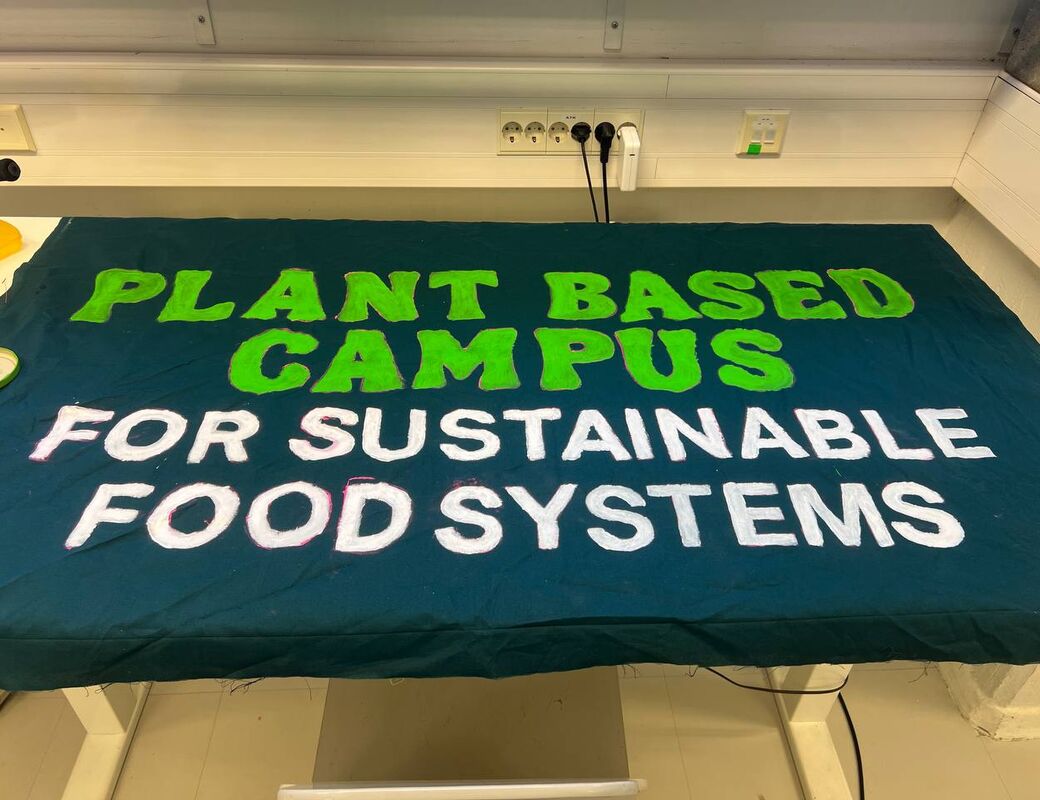 Hand-painted green and white banner on a table reading: PLANT BASED CAMPUS FOR SUSTAINABLE FOOD SYSTEMS