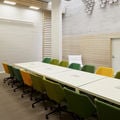 Long, white table surrounded by 20 chairs in light and dark green and yellow. Backdrop is white concrete wall with some birch cladding. Photo by Aalto University / Tuomas Uusheimo
