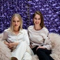 kaksi naista istuu vaalealla karvataljalla violeteista putkista koostuvan taustan edustalla ja katsoo kameraan