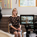 A picture of a video shoot, where a smiling woman is sitting in front of a video camera and a director is watching the scene.