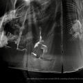 Image Description: black and white photograph of artist Yo-Yo Lin performing on a stage with right knee and her left hand touching the floor. She is surrounded by a translucent scrim with graphical images projected upon it.   the image credits:  the walls of my room are curved (2019). Courtesy of artist Yo-Yo Lin. Photo by Steve Dabal.