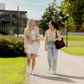 Two female students walking in a lush and green Aalto University campus, looking at each other while talking and walking.