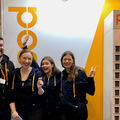 Four people are wearing dark hoodies, standing infront of orange wall with Posti logo, smiling and waving. One is showing a victory sign with fingers.
