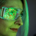A closeup of a woman in green light wearing goggles that reflect colourful pixels 