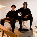 Two men dressed in dark clothes sit in the middle of furniture they have designed