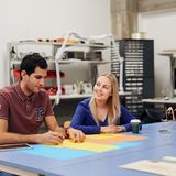 Two students drawing on coloured paper at a table in a workshop space