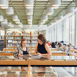 Kaksi opiskelijaa selailemassa lasisen vitriinin päällä olevaa kirjaa Oppimiskeskuksessa.