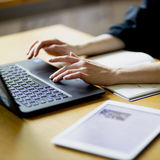 A person typing on a laptop, with notebook and table open, sun shines on the items