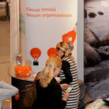 People in front of colorful roll-ups with pictures of rocks and Finnish text. People are talking to each other, some are reading books. Candy and computer on the tables.