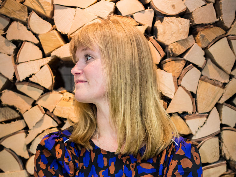 Anniina Suominen, an Associate Professor of Art Pedagogy at Aalto University’s School of Arts, Design and Architecture. She is standing in front of the camera, smiling, arms crossed. In the background there is a stack of logs.