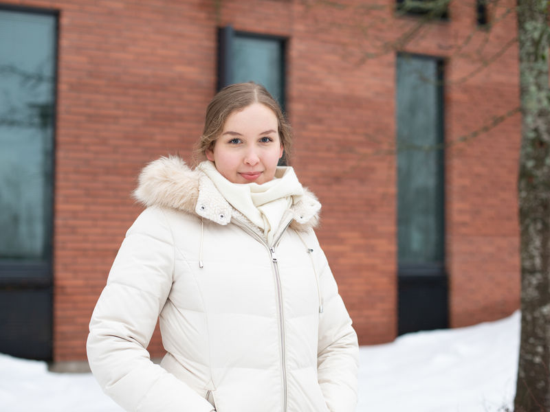 Heli Julkunen seisoo ulkona ja katsoo hymyillen kameraan. Taustalla näkyy oranssinruskea tiilirakennus ja lumihankia.