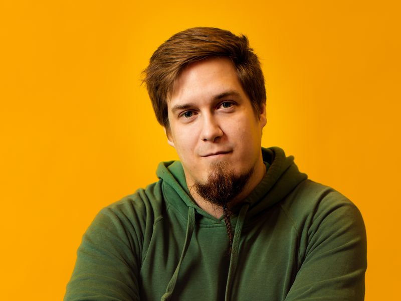 Picture of Simo Lahdenne in a darkgreen hoodie towards an orange background. He has crossed arms and is looking into the camera.