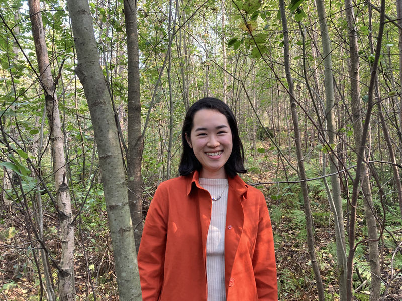 Mariko Yoshida in a red jacket standing in the forest, smiling directly into the camera.