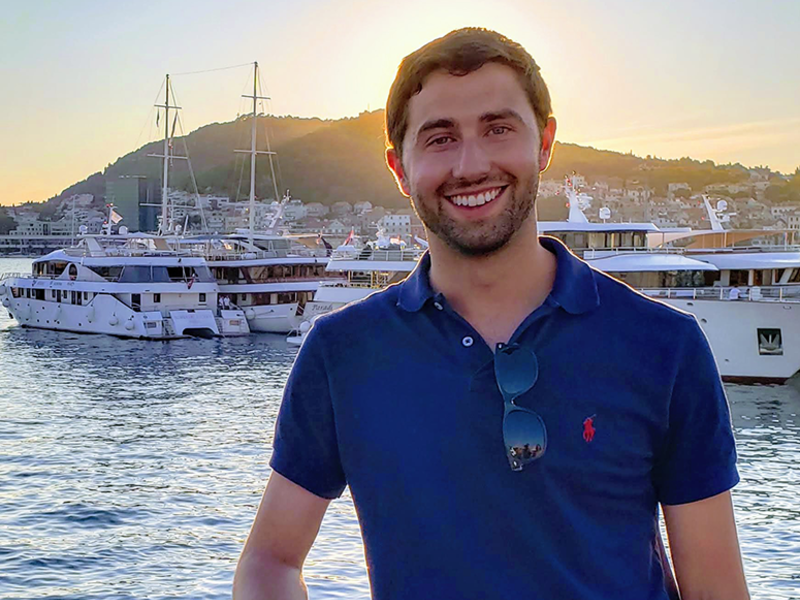 Tyler Spring in a blue shirt standing in a harbour, smiling towards the camera with the sea and boats in the background.