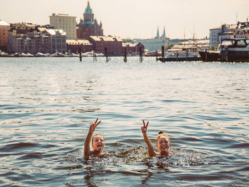 Two women swimming and showing the peace sign in front of an urban Helsinki city view. 