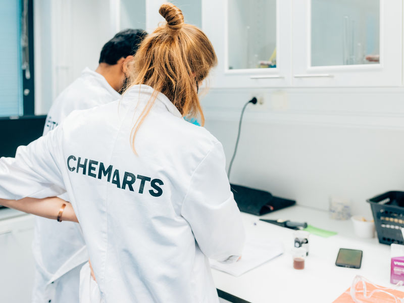 Two Aalto University Summer School students in lab coats with the text CHEMARTS on the back experimenting in the lab.