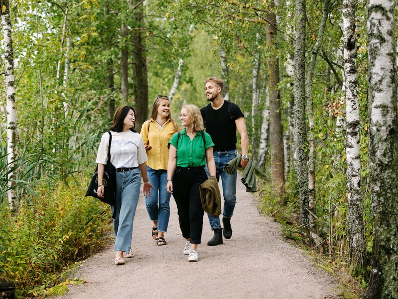 Four Aalto University Summer School students walking and chatting in the lush, green forest areas around Aalto University campus in the summer.