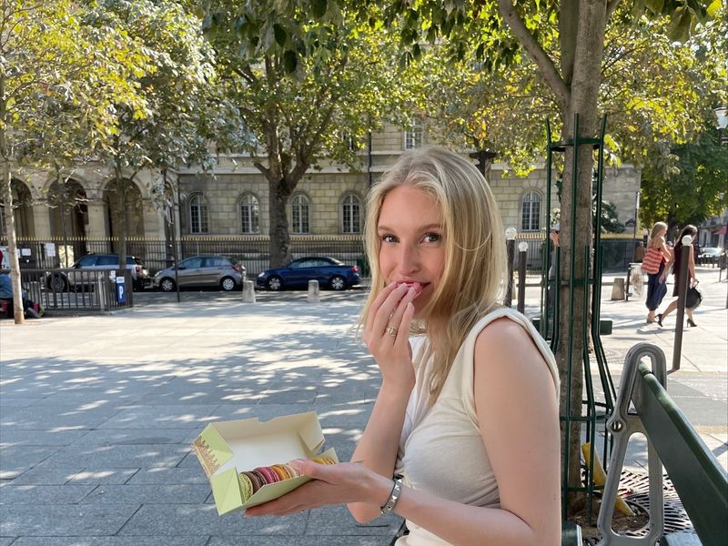 Kuvassa on kesä, ja Karla Helenius istuu puiston penkillä syömässä pieniä leivoksia. 