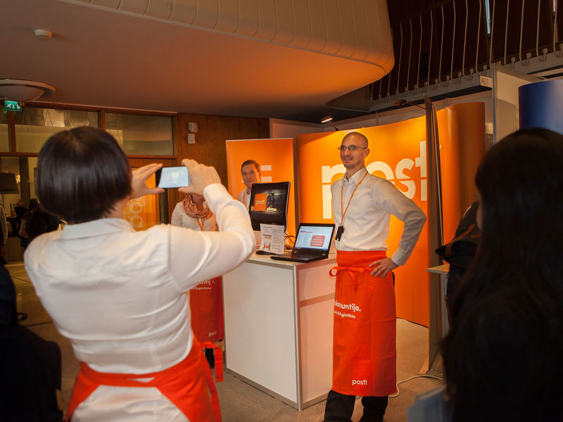 A person is standing with a back facing the camera, and holding a camera infront of their head. The person is shooting the camera at another person, who is standing next to a table with a laptop and infront of an orange expo walls with Posti logo.