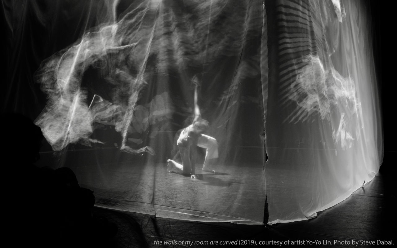 Image Description: black and white photograph of artist Yo-Yo Lin performing on a stage with right knee and her left hand touching the floor. She is surrounded by a translucent scrim with graphical images projected upon it.   the image credits:  the walls of my room are curved (2019). Courtesy of artist Yo-Yo Lin. Photo by Steve Dabal.