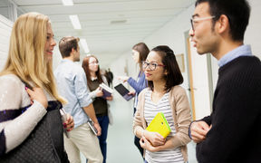 Several students, women and men discussing in the school hallway. One of the women is holding a yellow book.