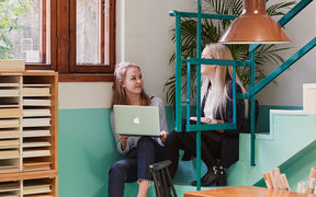Kaksi opiskelijaa istumassa paperikaupan värikkäillä rappusilla kannettavan tietokoneen kanssa.