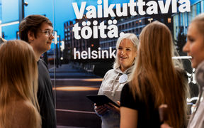 A blond woman is holding an ipad and talking to students infront of a colorful wall with text Vaikuttavia töitä