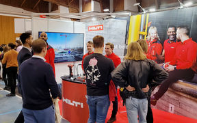 People are standing in front of a wall with huge picture of people with red shirts sitting on a coffee break and a Hilti logo. People are talking with eachoterh.
