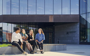 Four people sitting on a bench outside a Building with large glass windows and a School of Business logo. People are laughing.