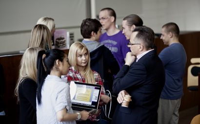 Two business students, one is holding a laptop, discussing with the Professor after his lecture. Another group of students talk in the background.