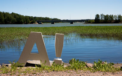 A white, 80 cms high 3D logo with capital A and an exclamation mark to the right of it. The logo is standing in the sunshine on a beach with the sea and some seagrass behind it. Picture by Aalto University / Mikko Raskinen 