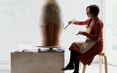 Professor Maarit Mäkelä sits on a chair and paints a ceramic pot with soil from Venice, in a brightly lit area