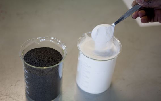 Kuvassa on kaksi mittalasia, joista toisessa on tummaa teräskuonaa ja toisessa valkoista kalsiumkarbonaattia. Kuvaaja: Mikko Raskinen.