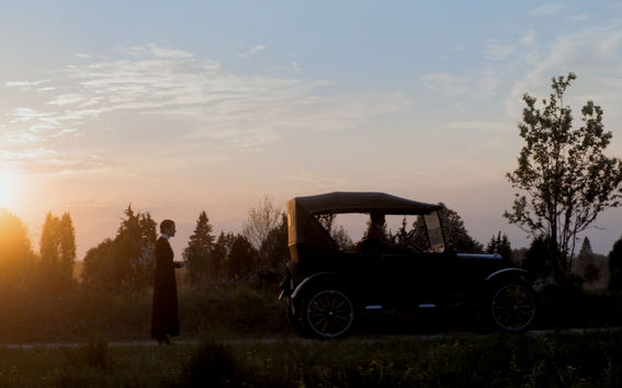 Helene-elokuvan kohtauksessa näkyy laskevan auringon maisemassa vanhanaikainen auto ja sen takana nainen seisomassa pää hieman painuksissa.