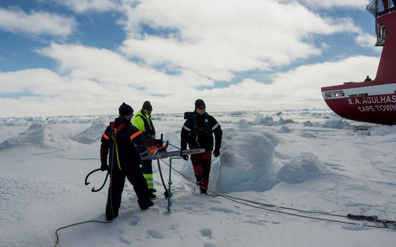 Kuvassa kolme miestä seisoo jäällä, yksi pitelee moottorisahaa. Taustalla näkyy punainen tutkimusalus.