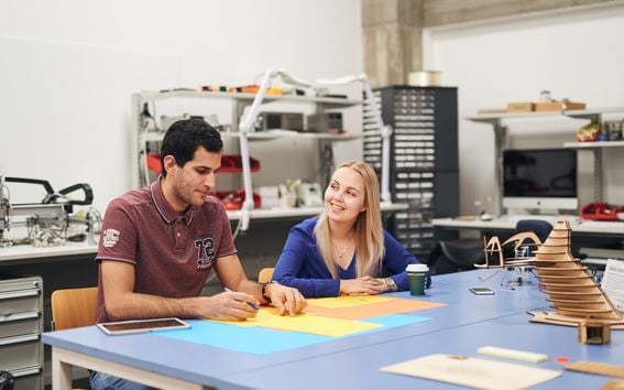 Two students drawing on coloured paper at a table in a workshop space