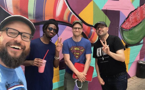 Four smiling men posing in front of a colorful graffiti wall. 