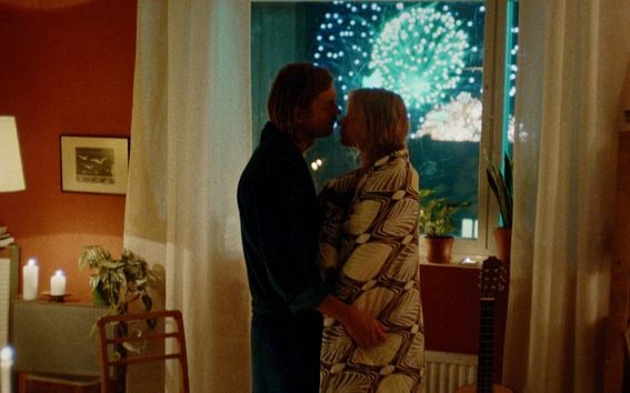 Still image from movie with a man and a woman embracing in front of a window displaying fireworks