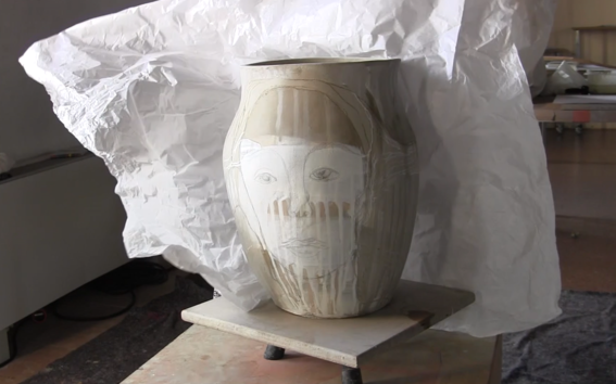a ceramic vase with a face painted on it. behind a wrinkled white paper