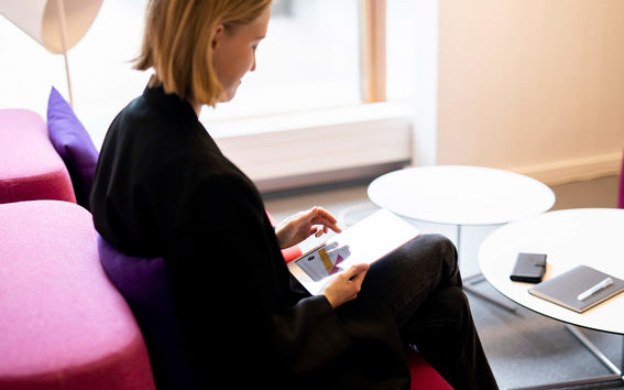 Toimistossa istuva nainen selaa tablettiaan, pöydällä esillä puhelin, muistikirja ja kynä.