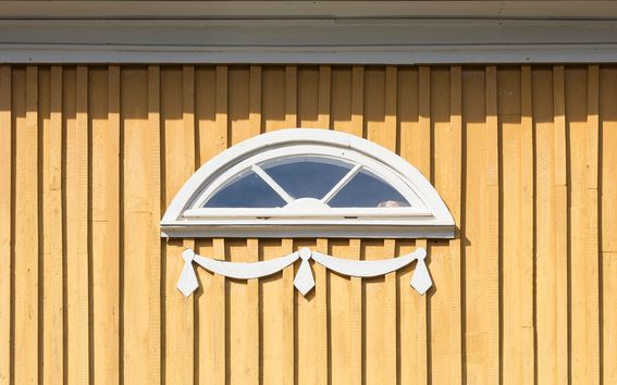 Yksityiskohta: keltaisen puurakennuksen puolikaaren muotoinen ikkuna valkoisine karmeineen ja koristeluineen