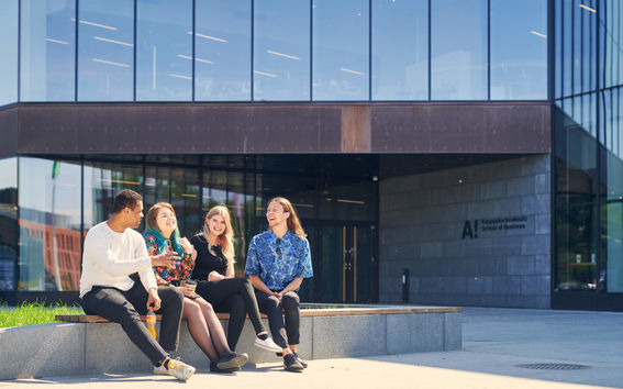 Four students sitting in on a wall outside the Aalto University School of Business building. They are all laughing and discussing vividly.