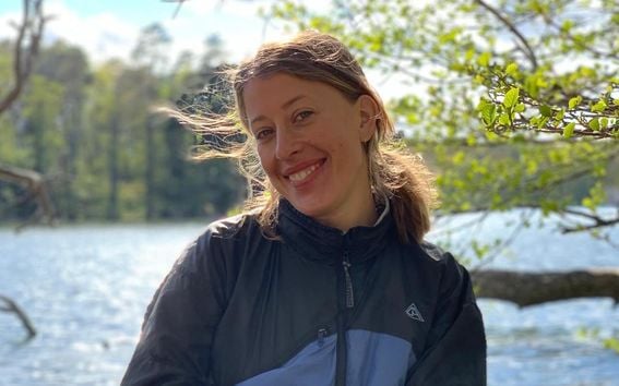 Carolina Weigl alumna from Aalto University Summer School course Nordic Biomaterials with CHEMARTS sitting in the nature by a lake in an outdoor jacket smiling directly into the camera.