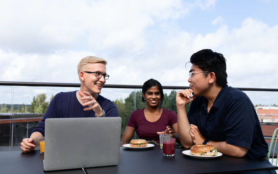 Three students eating a snack and discussing over a computer on a rooftop terrace. 