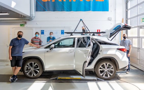 Autonominen A!ex-auto kuvattuna valoisassa autohallissa ympärillään viisi tutkijaa kesäisissä vaatteissa ja kasvoillaan maskit. Valkoisen auton katolla on tekninen laite ja auton tavaratilan kansi on ylhäällä.