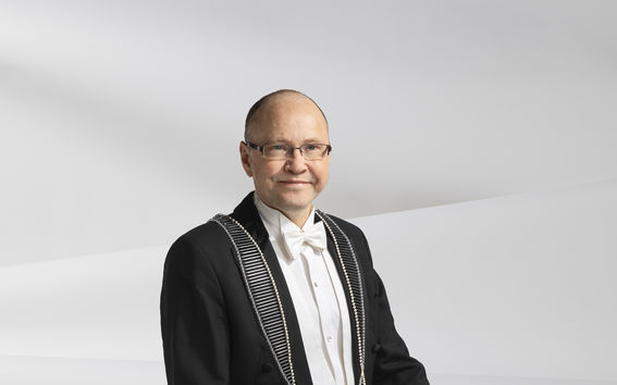 Janne Laine sits on a tall black chair, dressed in a tuxedo, wearing the Dean of School of Cheminal Engineering ceremonial link necklace, smiling. Background is white and slightly wavy.