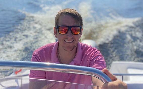 Matthew Clarkson in a pink shirt and mirrored sunglasses pictured driving a boat on a sunny day at the sea.